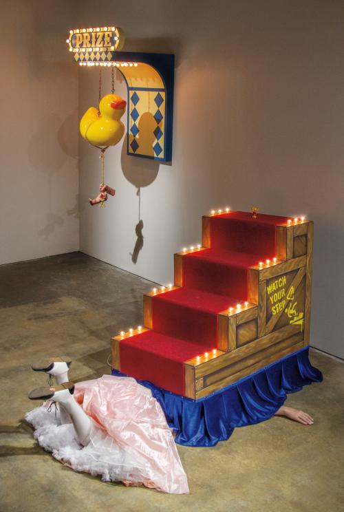 Photograph of an art installation featuring a person lying on the floor partially covered by a red-carpeted staircase with lit candles along edges and a "Watch Your Step" warning on the side. Above, a large yellow rubber duck hangs from a sign labeled "Prize," with a blue and yellow geometric wall decoration in the background.