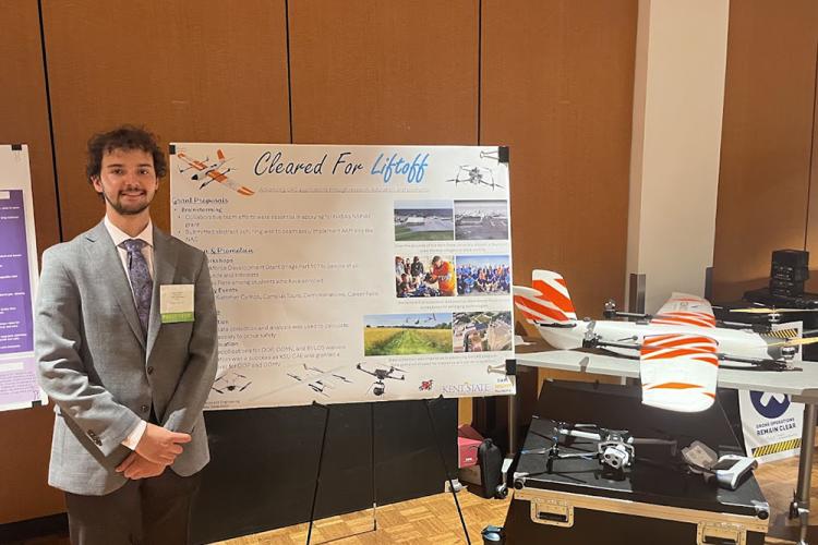 A young man in a grey suit stands next to a poster and a flying drone aircraft.