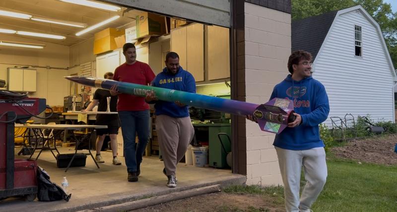 Three men are carrying a long rocket out of a garage
