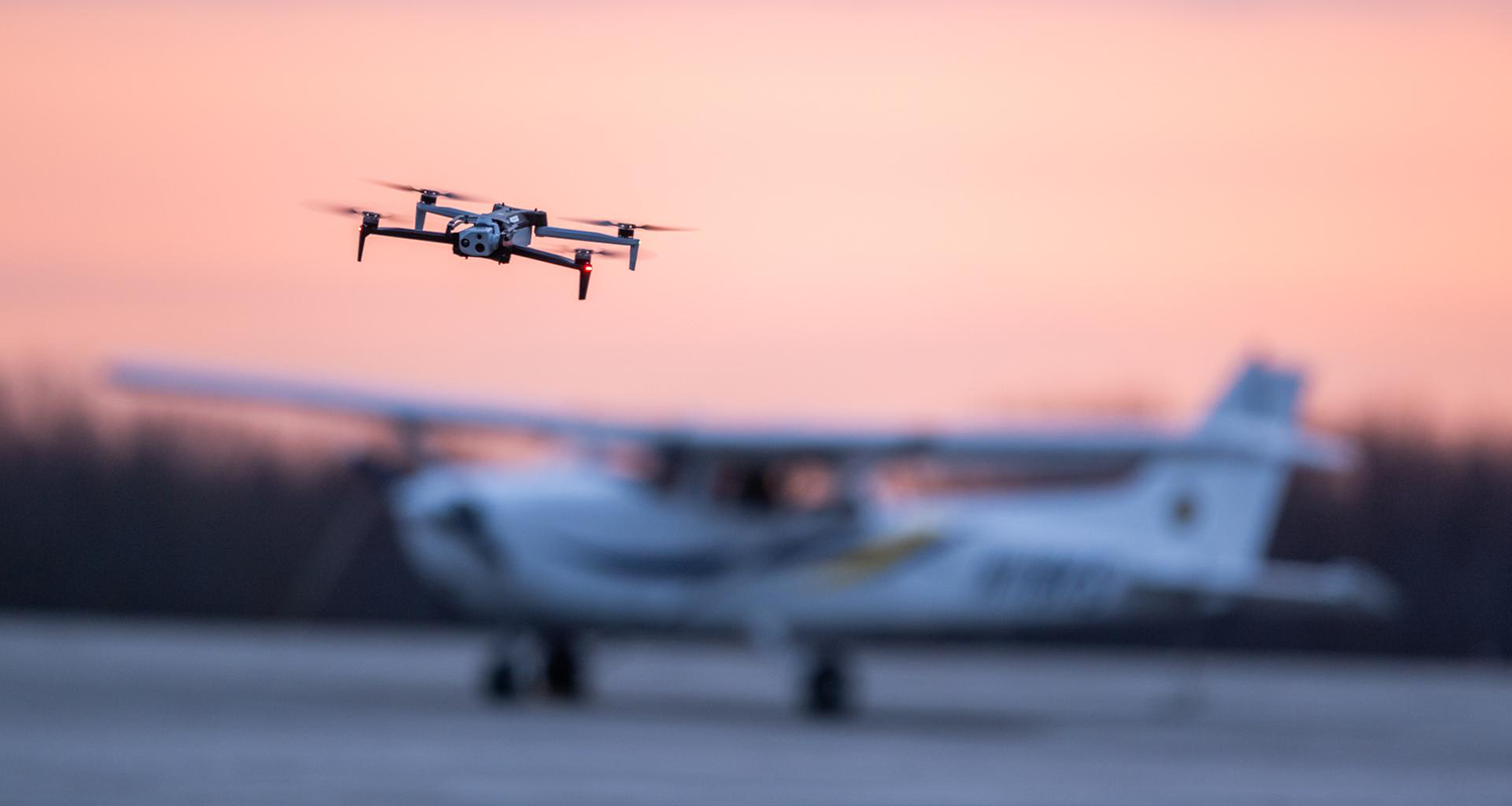 A quadcopter drone flying in the forground with a slightly out of focus small airplane in the background.