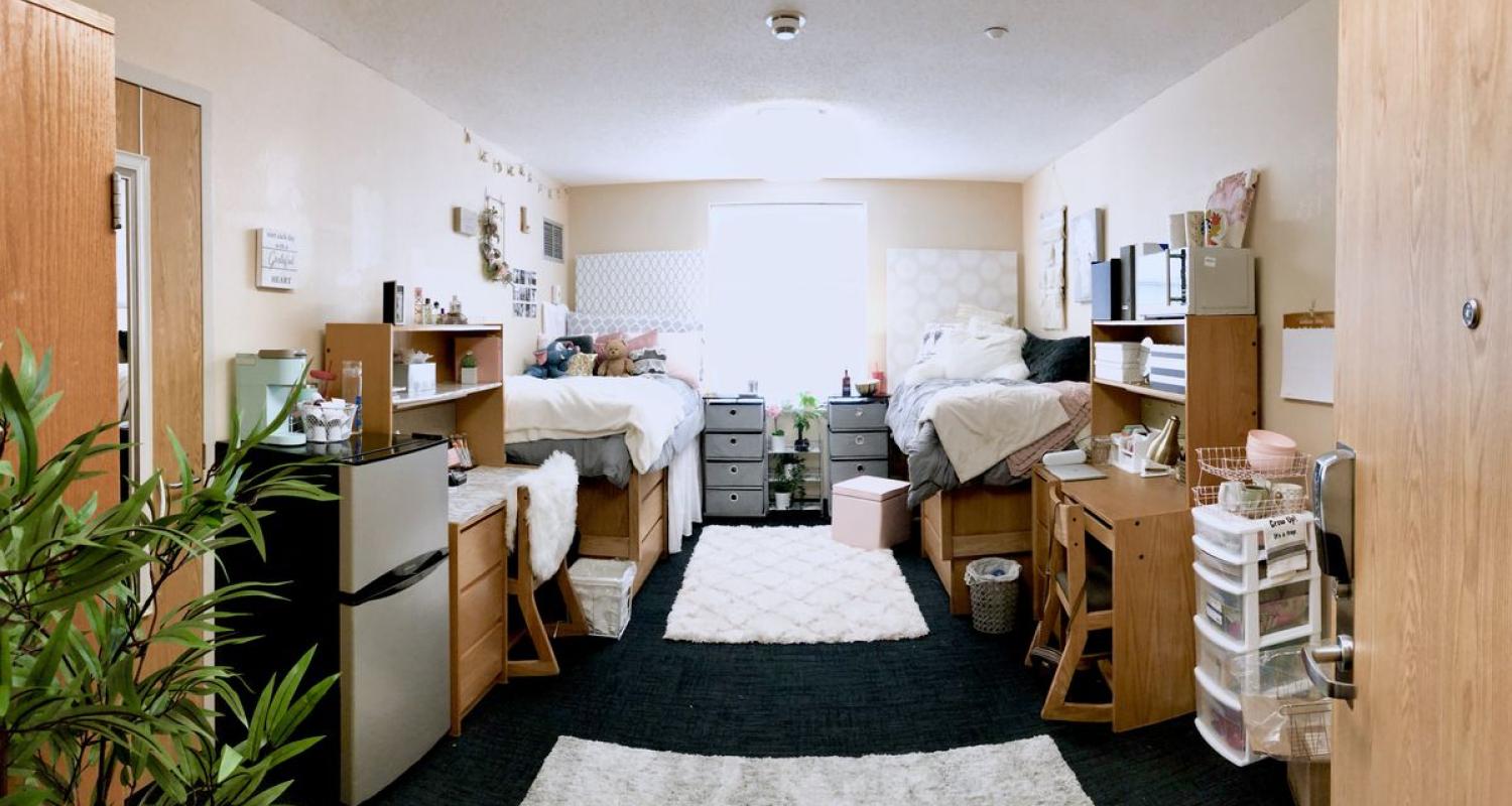 A room with two beds with coordinating white and grey bedding, pink accessories, a white rug and plants