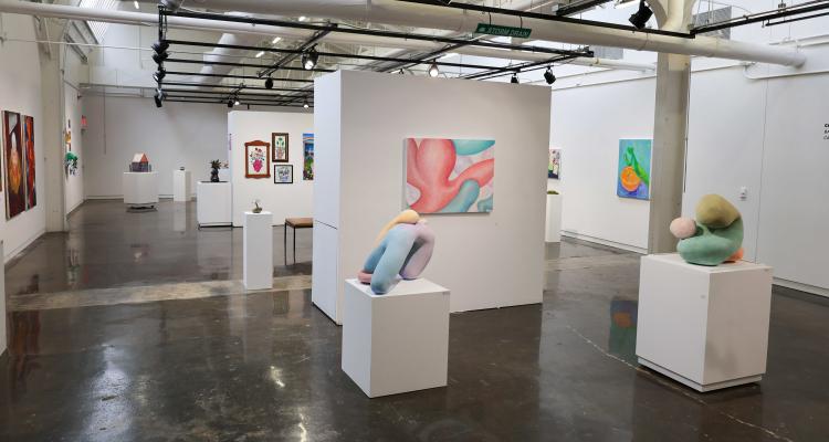 Wide view of a contemporary art gallery with polished concrete floors, white walls, and track lighting, featuring colorful abstract paintings on the walls and soft, organic-shaped sculptures displayed on white pedestals throughout the open space.