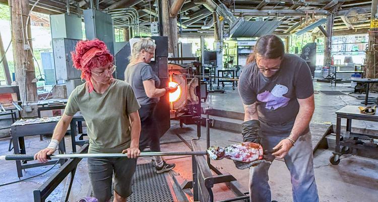 Wide view inside a glassblowing studio with a high wooden-beam ceiling and industrial equipment, showing multiple people working at separate stations; in the foreground, two people shape a glowing molten glass piece on a metal rod at a workbench covered with tools, while additional workstations and a furnace are visible in the background.