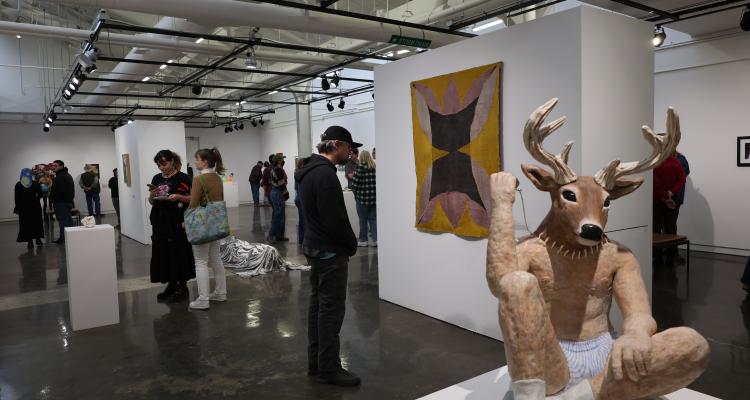 Gallery with people looking at art featuring sculpture of a deer head on human body in foreground