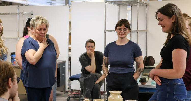 An artist smiles down at her body of work, including an assortment of ceramic mugs and pots, while a crowd smiles and studies it