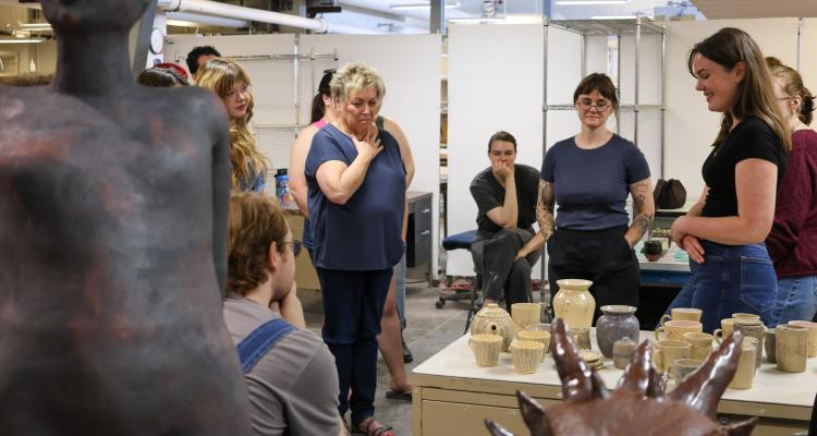 A smiling student stands before an assortment of ceramic mugs and containers as a small crowd gathers to critique her work