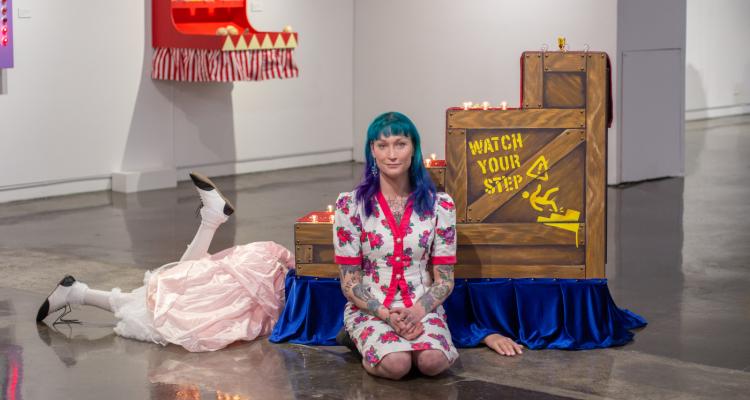 Violet kneels alongside two of her exhibition pieces. Behind her is a staircase reading "watch your step" with legs kicking out from beneath.