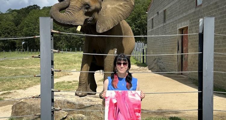 Natalie Holland standing in front of the elephant Sukiri holding a painting.