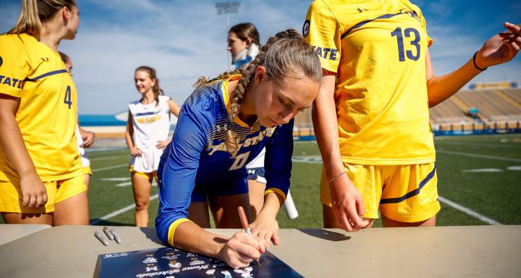 Emma Klein signing a poster on the field while surrounded by fellow team mates. 
