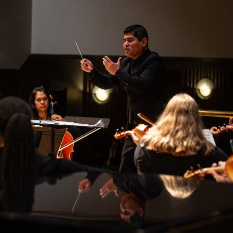 Conductor leading a group of student musicians playing string instruments, including violins, cellos, and a double base.