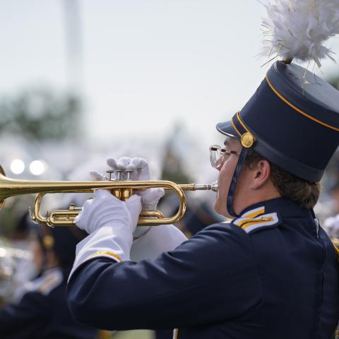 Marching band musician in navy uniform and tall shako with white plume plays trumpet during an outdoor performance, with other band members blurred behind.