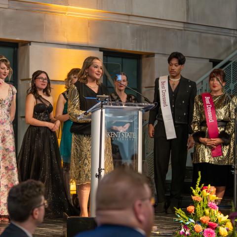 Museum director gives a speech at the dazzle gala while students stand behind her wearing best dressed sashes