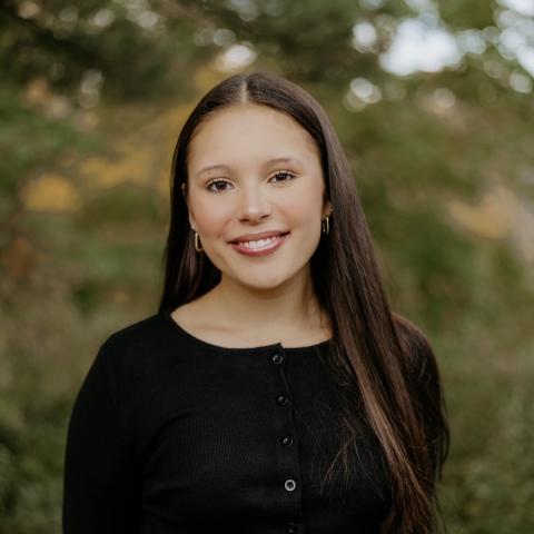 Audrey in a nice black blouse in an outdoor setting smiling with her mouth closed while looking at the camera and some of her long brown hair blowing behind her. 
