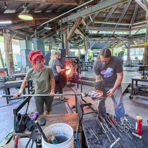 Wide view inside a glassblowing studio with a high wooden-beam ceiling and industrial equipment, showing multiple people working at separate stations; in the foreground, two people shape a glowing molten glass piece on a metal rod at a workbench covered with tools, while additional workstations and a furnace are visible in the background.