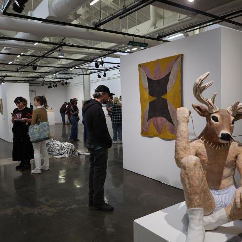Gallery with people looking at art featuring sculpture of a deer head on human body in foreground
