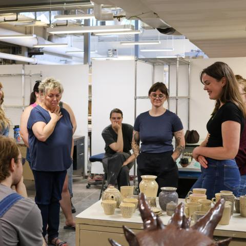 An artist smiles down at her body of work, including an assortment of ceramic mugs and pots, while a crowd smiles and studies it