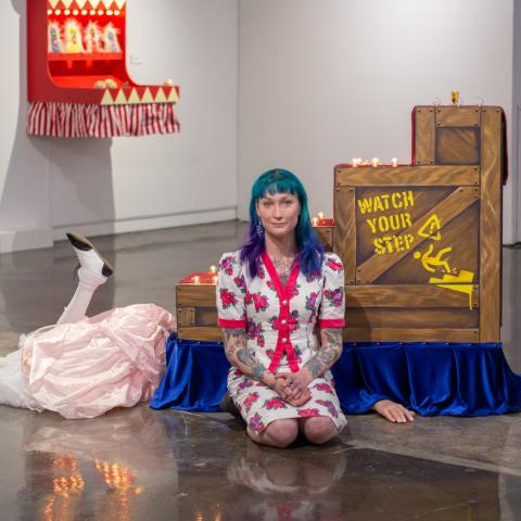 Violet kneels alongside two of her exhibition pieces. Behind her is a staircase reading "watch your step" with legs kicking out from beneath.