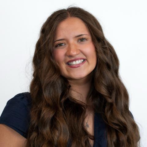 Professional headshot of Noelle with long, wavy brown hair and a large smile while wearing a dark blue blouse against a white background. 
