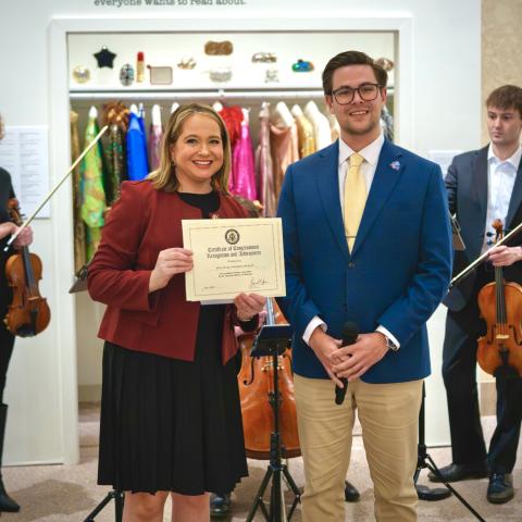 Two adults stand indoors holding a framed “Certificate of Congressional Recognition and Achievement” while several musicians with string instruments stand behind them; one person holds a microphone. The background shows a display of colorful garments or costumes on racks within a museum or gallery setting.