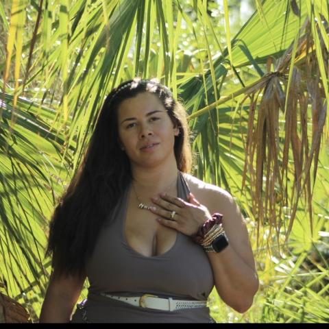 Waist up shot of Siera standing against background of palm trees. She has long brown hair and wears an army green tank top