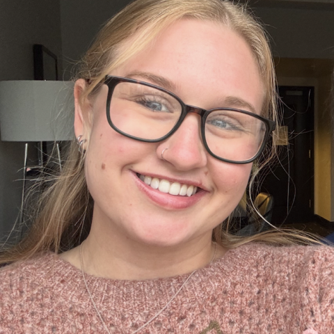 Taylor in a light pink sweater with a silver necklace smiling in a selfie with large tortoiseshell patterned glasses.