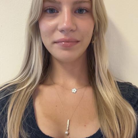 Bella in a long sleeve ribbed blue shirt softly smiling at the camera in a selfie against a blank wall, with a light golden necklace on her neck and long blonde hair on her shoulders. 