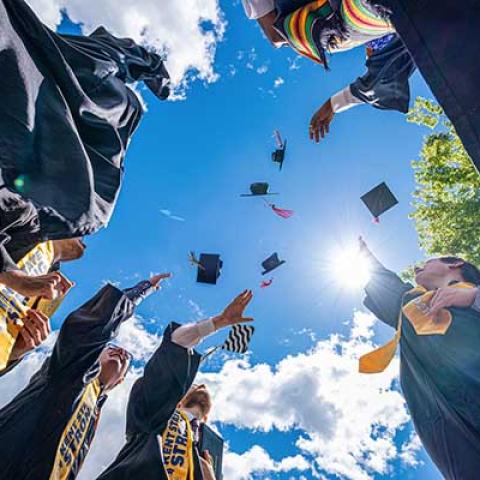A view straight up in a circle of graduates as they toss their caps into the air against a blue sky