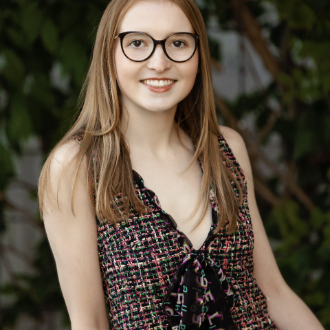 Olivia leaning on one arm in a patterned tank top smiling at the camera with large black glasses on her face.