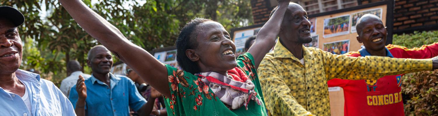 A group of adults dance and celebrate outdoors near a community building, arms outstretched and smiling, with a bulletin board displaying photos visible in the background.