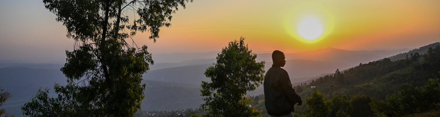 A lone figure stands on a hilltop road at sunset overlooking a wide valley in Rwanda, with eucalyptus trees framing a golden horizon and rolling hills receding into the distance.