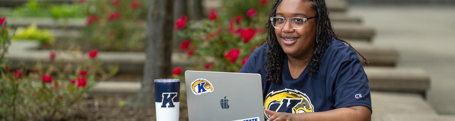 Student in ent State shirt sitting at a table outside with a laptop