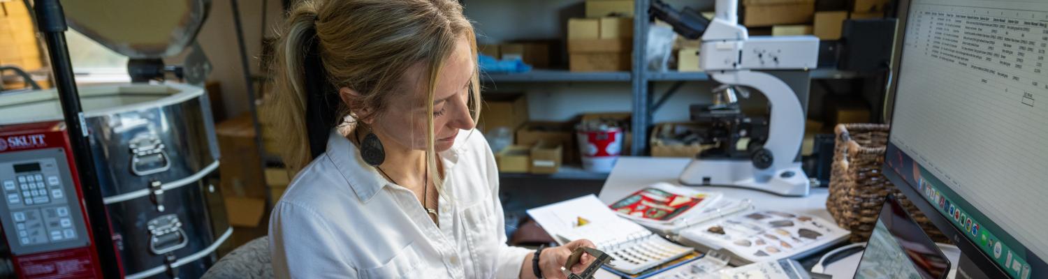 Dr. Michelle Bebber working at a desk in an archaeology lab, carefully measuring a rock. 