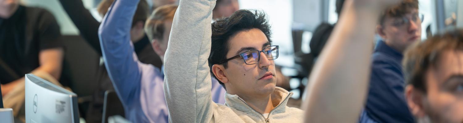 A student wearing a gray sweatshirt and glasses raises their hand in class, with other students around them also participating.