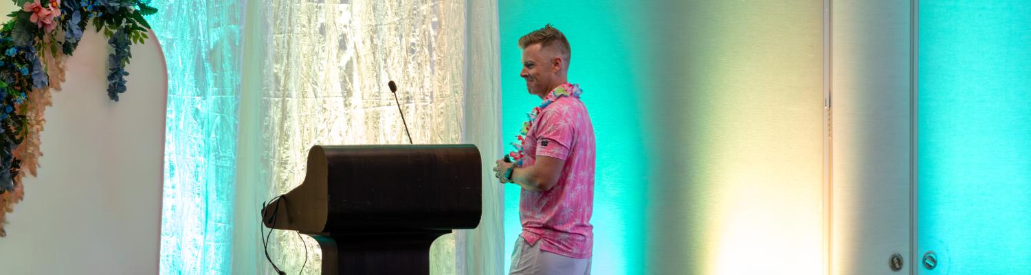 Ben Lorenzen smiles while speaking at a podium during the 2026 Schwebel Lecture. He is wearing a pink tropical-print shirt, khaki shorts, and a blue floral lei. The stage is decorated with a tropical theme, featuring teal lighting, a beach ball, and floral accents. Several audience members are visible in the foreground.