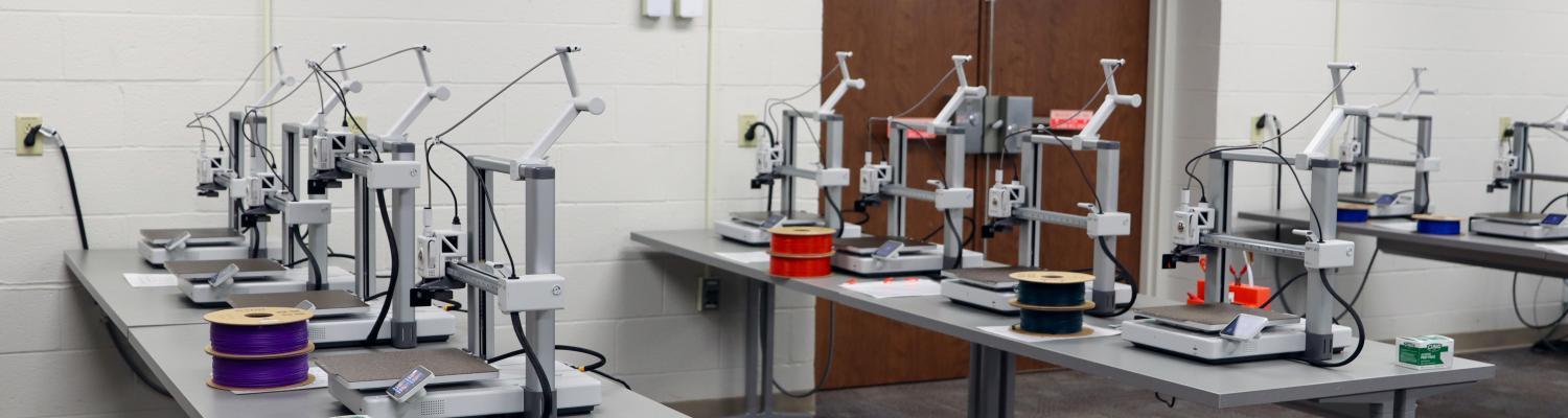 A classroom with multiple 3D printers arranged on gray tables in rows. Each printer has a spool of filament in different colors, such as purple, red, and green. The room has white walls, a carpeted floor, and double wooden doors in the back.