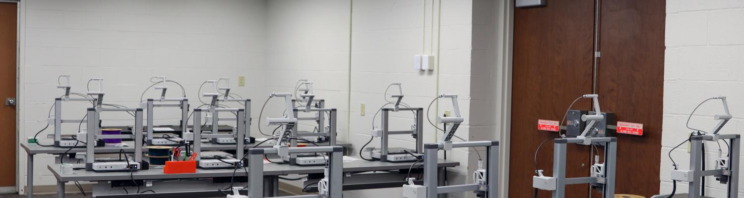 A classroom setup with multiple 3D printers arranged on gray tables in rows. Several printers have filament spools attached, including yellow, purple, blue, and green. The room has white walls, carpeted flooring, fluorescent ceiling lights, and double wooden doors at the back.