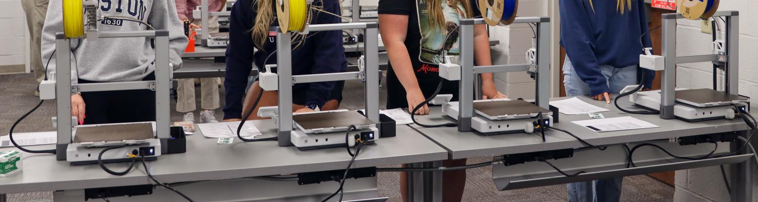 A group of students works in a classroom with several 3D printers lined up on tables. The printers have spools of yellow and blue filament loaded. Four students in the foreground are standing at the machines, reading instructions or adjusting settings, while more students and an instructor are visible in the background. The room has white walls, fluorescent lighting, and wooden double doors.
