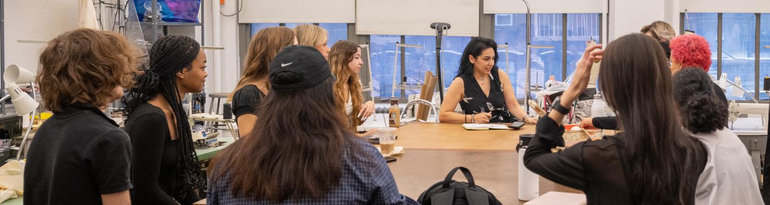 Students gather around a square table, directing their gazes at a woman with dark hair
