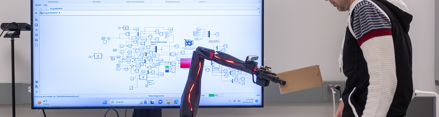 A male student observes a small robotic arm completing a task of lifting items and stacking them on a table