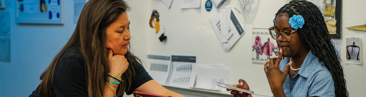 A woman sits with another, pointing to a card on the table between them. 