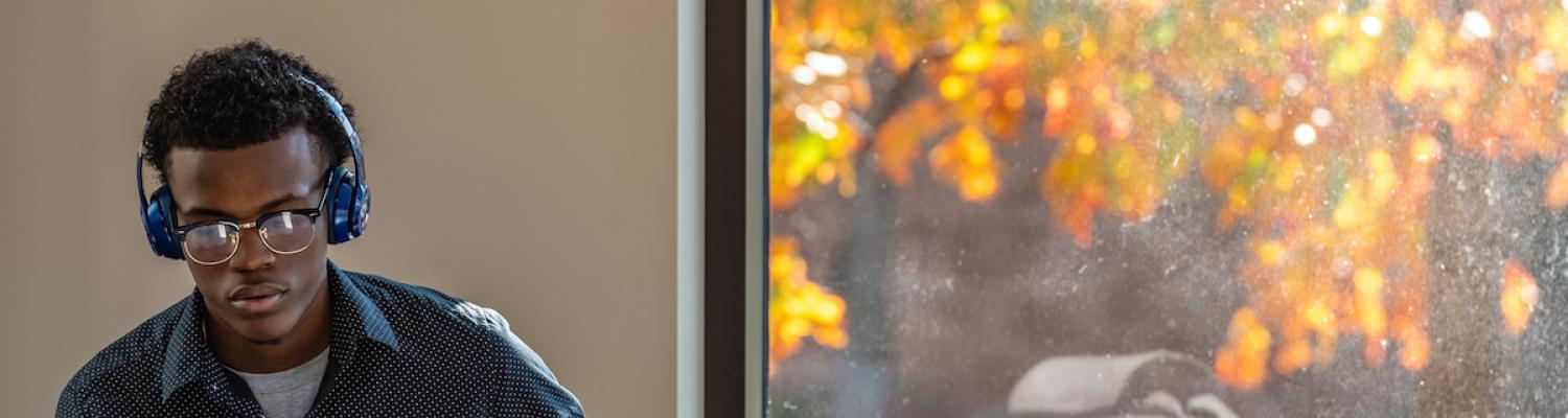 Male student sitting by the window studying on the computer with headphones on.