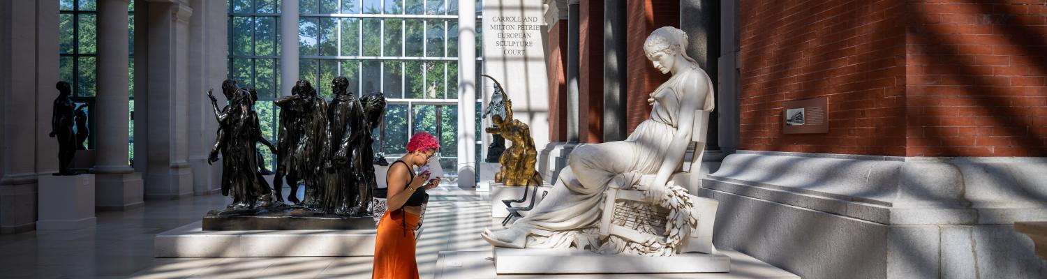 A student in an orange skirt studies a white marble sculpture of a humanoid figure
