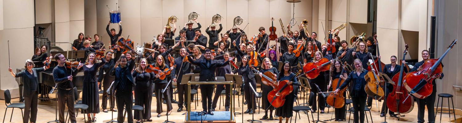 A large student orchestra poses onstage with string, brass, woodwind and percussion instruments, raising bows and instruments in celebration beneath red theater curtains.