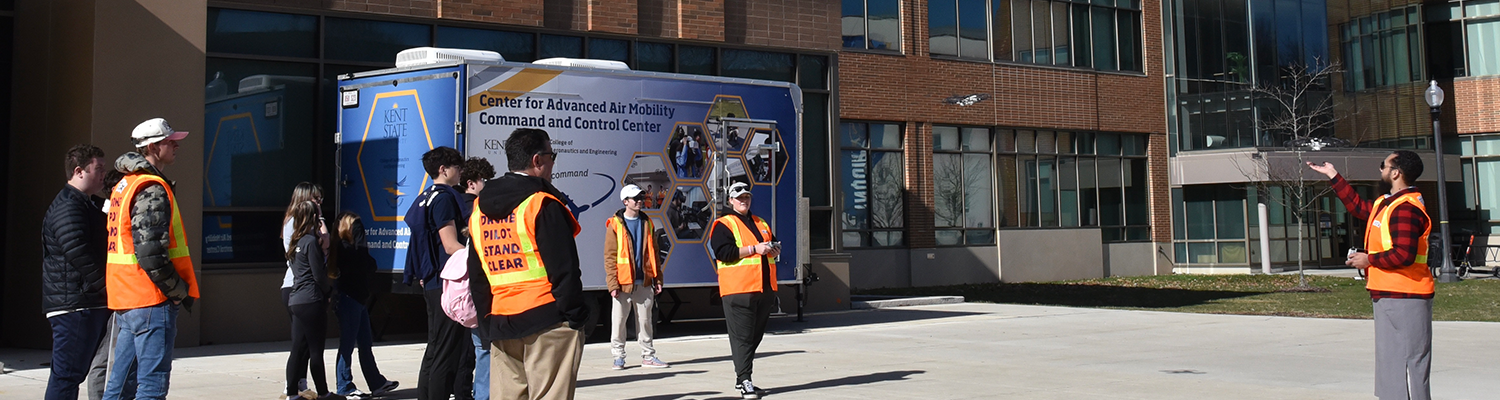 A group of students wearing orange safety vests operate drones in front of the Mobile Command and Control Center located in front of the Aeronautics and Engineering Building