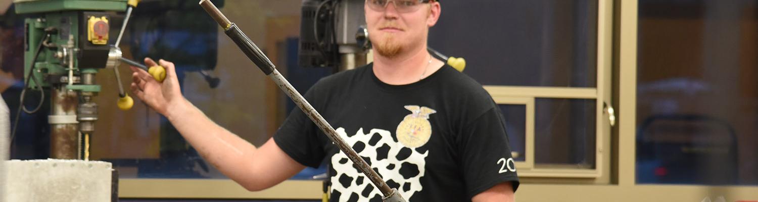 A male student wearing safety goggles stands by a drill press in the Materials and Processes lab