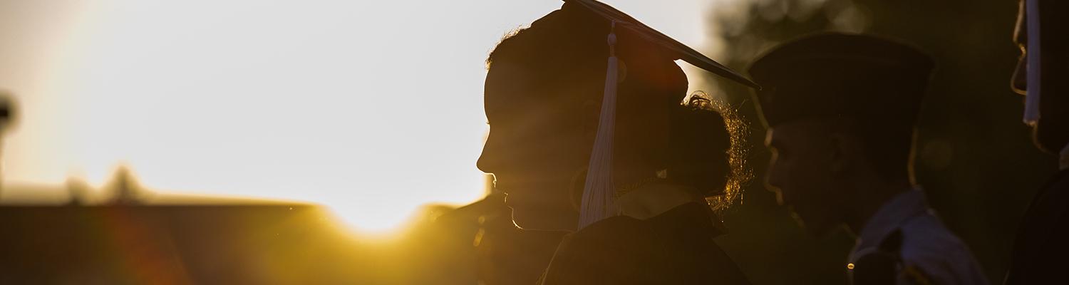 A picture of a young woman in a graduate cap with the sun setting behind her