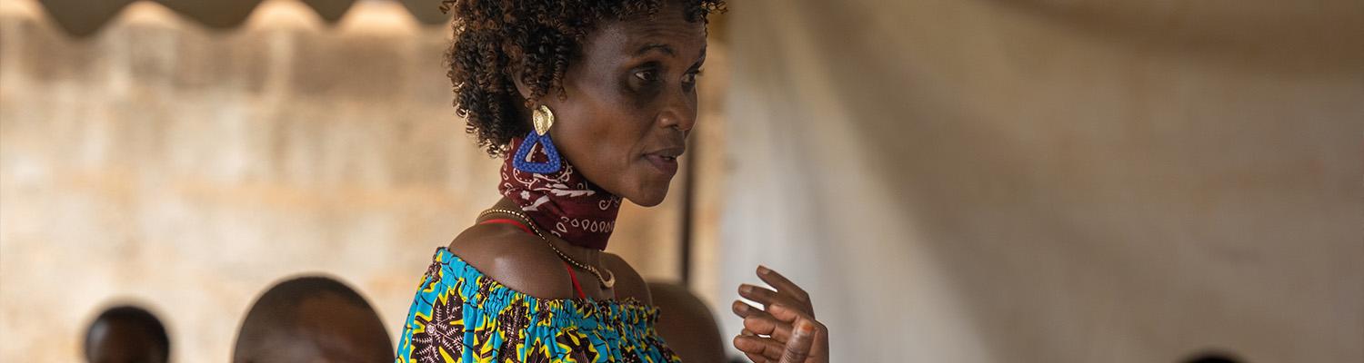 A woman in a vibrant ankara print off-shoulder top speaks and gestures to attendees inside an open-air tent, with seated participants listening in the background.