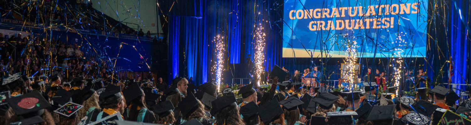 Large group of graduates in darkened room with screen reading "Congratulations Graduates"