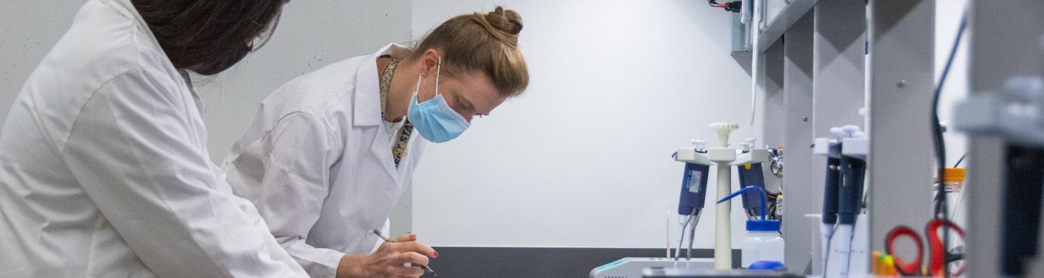 Two individuals in lab coats and one wearing a face mask work at a laboratory bench, using tools and writing notes beside scientific equipment.