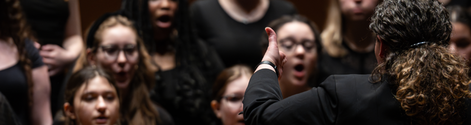 A group of students singing is blurred out in the background as a conductor leads them in the foreground